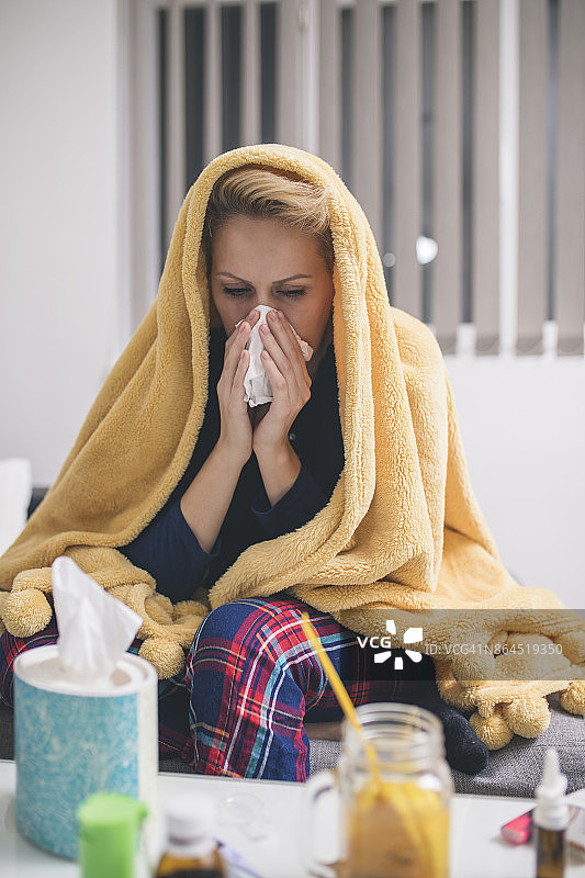 一个生病的年轻女子因感冒坐在家里的沙发上，她盖着一条毯子，擤着鼻子图片素材