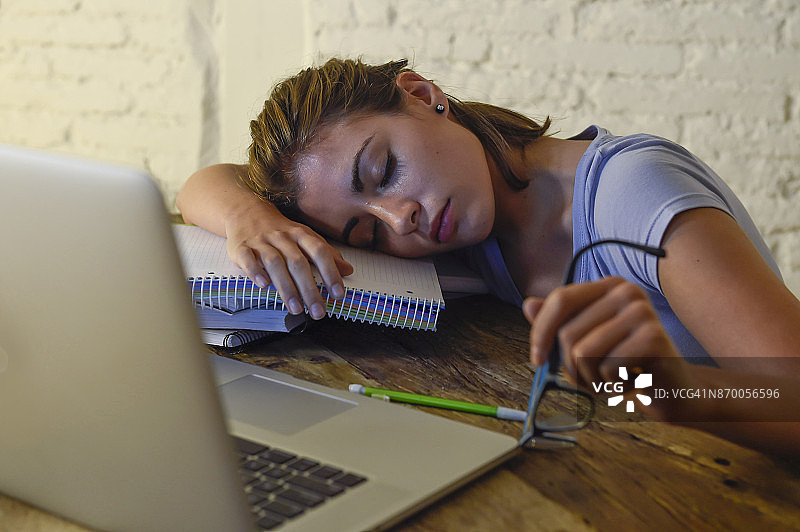 年轻美丽而疲惫的女学生躺在家里的笔记本电脑桌上打盹，精疲力尽，浪费了一个晚上在压力下学习准备考试图片素材