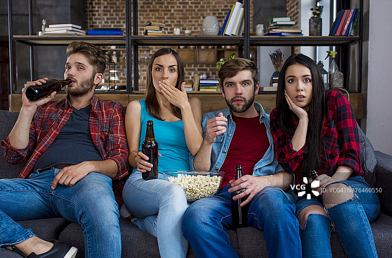 一群现代学生朋友在电视上看恐怖电影，坐在家里的沙发上喝着啤酒吃着爆米花。图片素材
