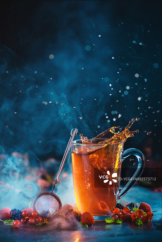 一杯热气腾腾的莓茶，用滤茶器和动态的飞溅，散落的茶滴和美丽的散景在一个黑暗的背景。各种新鲜浆果和薄荷叶。图片素材