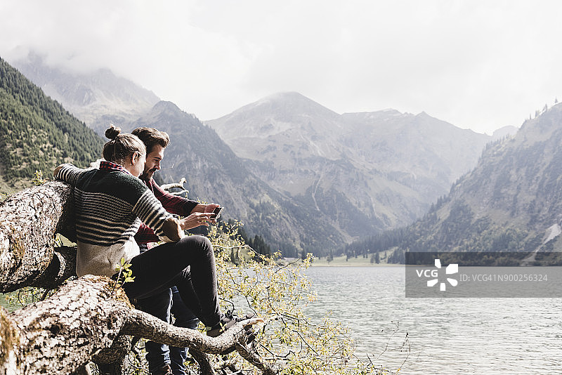奥地利，蒂罗尔，阿尔卑斯山，夫妇放松在树干上的山湖使用手机图片素材