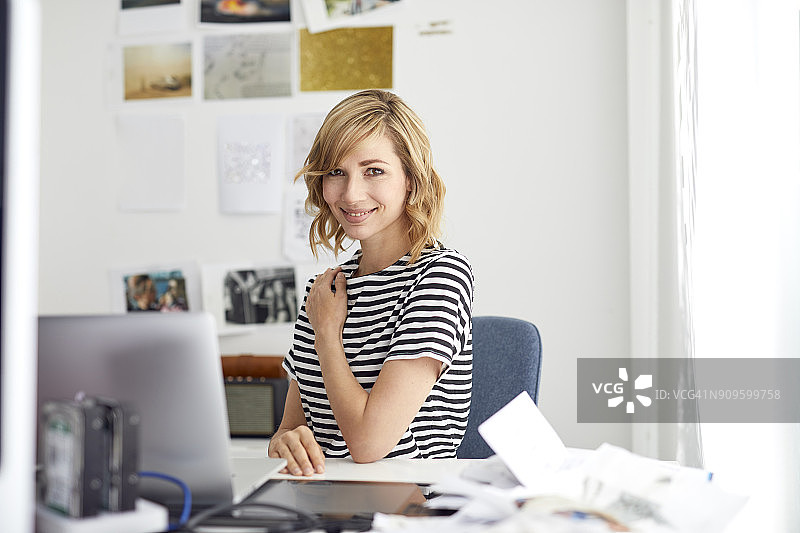 微笑的金发商务女性的肖像，手提电脑，坐在办公桌前图片素材
