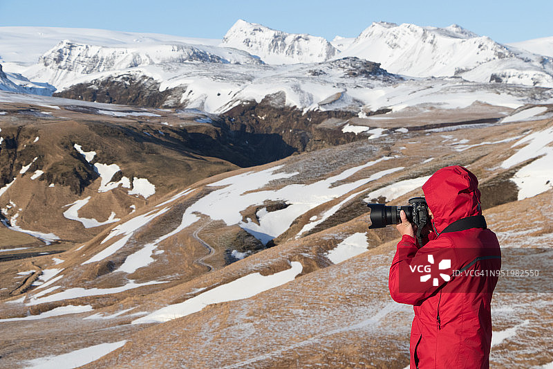 在前往冰岛南部维克市的路上，一名身穿红色夹克的男摄影师在视点拍摄雪山图片素材