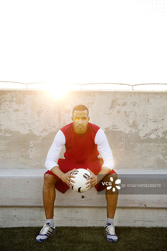 在阳光明媚的日子里，一个自信的运动员抱着足球坐在靠墙的座位上图片素材