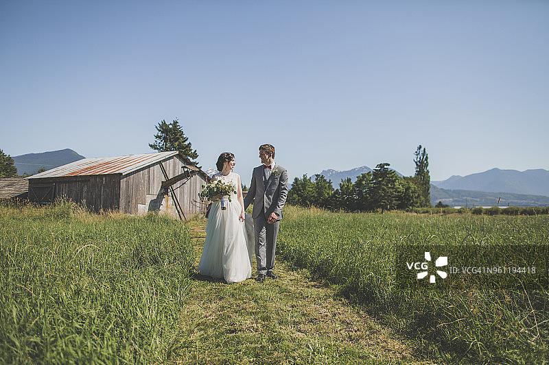 一对新婚夫妇在谷仓和晴朗的天空下的草地上面对面地走着图片素材