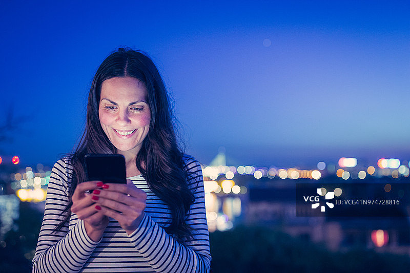 美丽的年轻女子使用手机对抗夜晚的城市景观图片素材