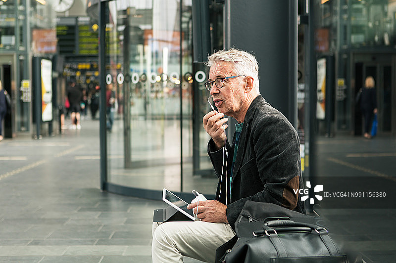在火车站里，坐在包边，用耳塞耳机说话的年长男性通勤者图片素材