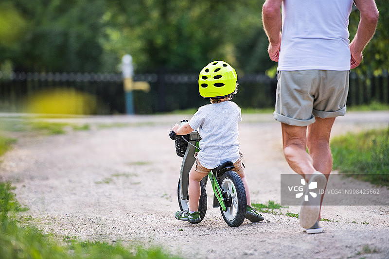 一个蹒跚学步的小男孩和他的祖父在户外骑着平衡自行车。后面的观点。图片素材