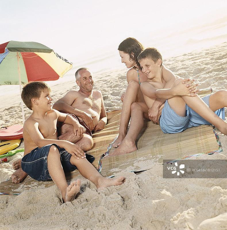 父母和他们的儿子坐在沙滩上的垫子上，用阳伞图片素材