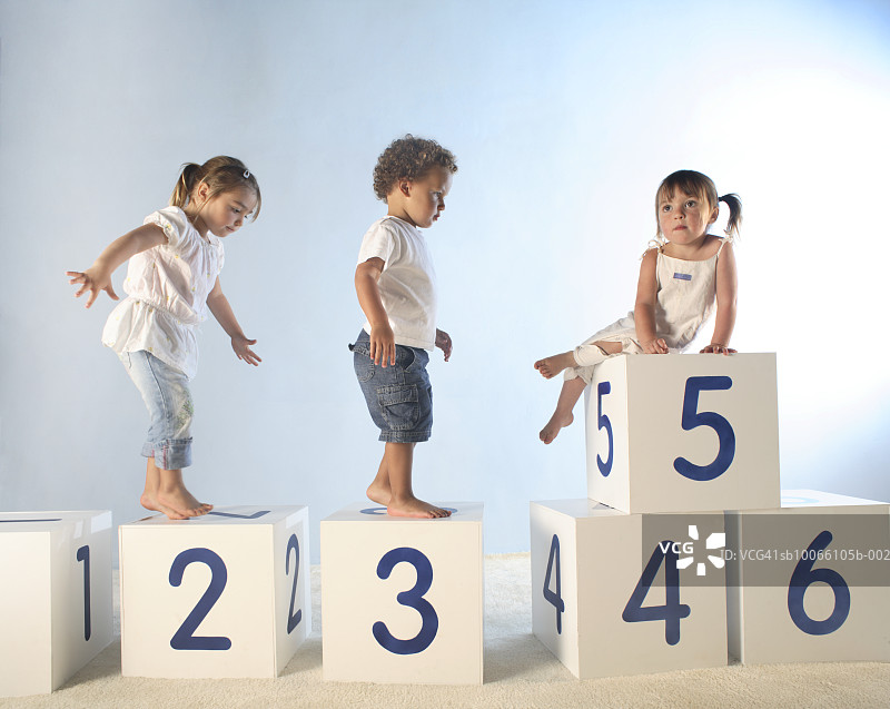 一个男婴(21-24个月)和两个女婴(2-5个月)正在爬上白色的字母盒子图片素材