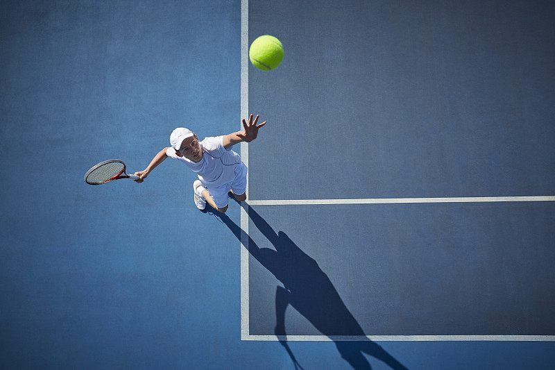 头顶上的年轻男子网球运动员在阳光明媚的蓝色网球场上打网球，发球图片下载