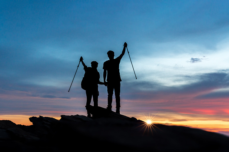 在日落时分，一对年轻的徒步旅行者正在攀登岩石山的顶峰，团队合作，帮助，成功，赢家和领导的概念。图片下载