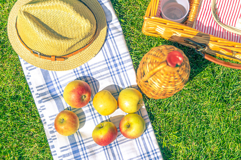 在花园里野餐，用老式的柳条瓶和柳条野餐篮，桌上铺着苹果和草帽图片素材