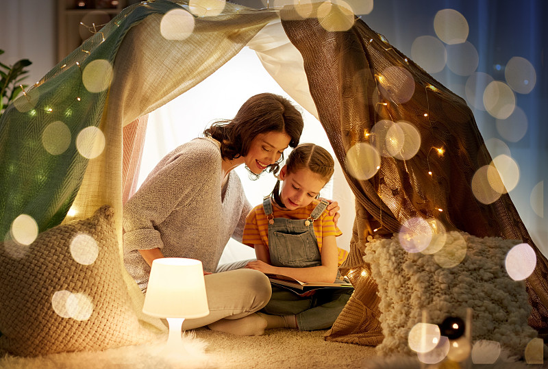 家庭、舒适和人的概念——快乐的母亲和小女儿晚上在家里的孩子帐篷里看书。快乐的家庭在家里的孩子帐篷里看书图片下载