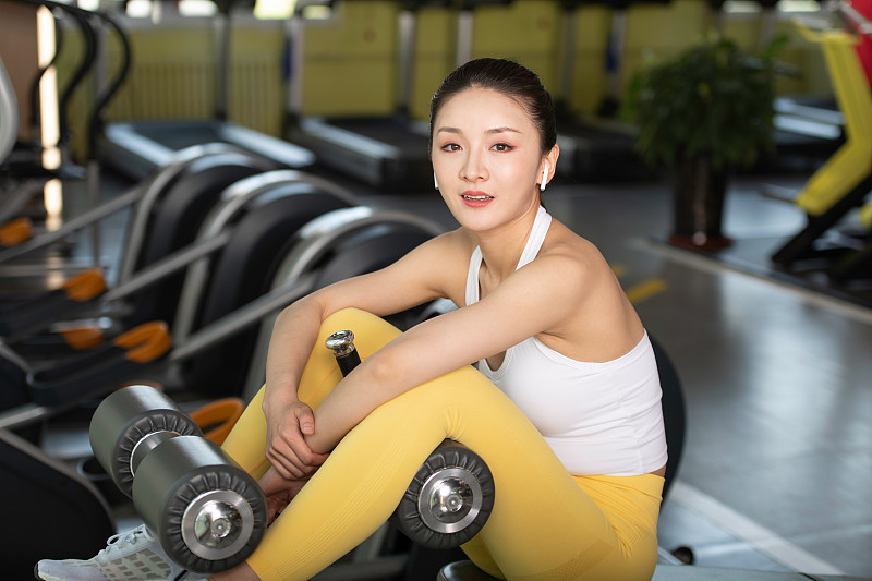 健身的年轻女性，在健身房训练间隙，坐在健身器材上休息。身材健美的年轻女人图片下载