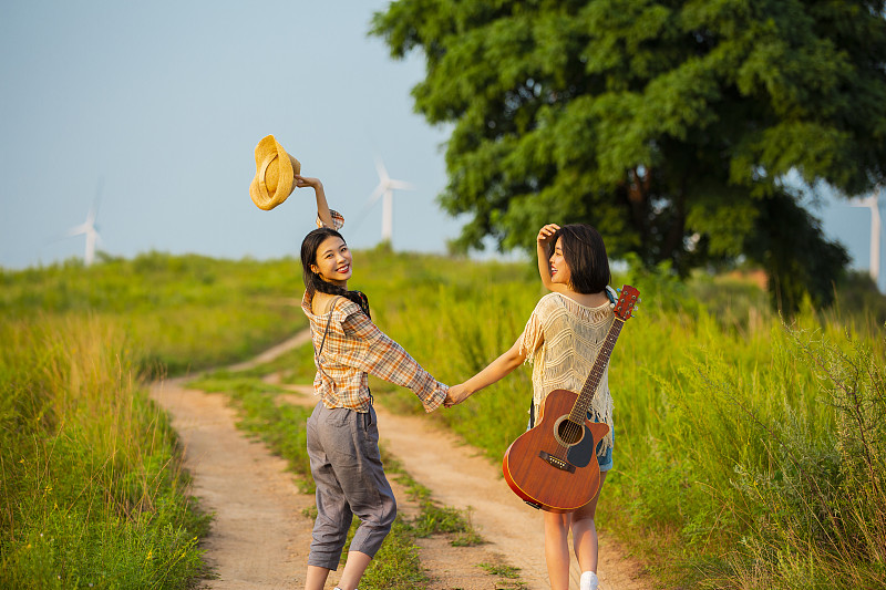 年轻闺蜜户外郊游，背着吉他手牵手走在乡村的小路上图片下载