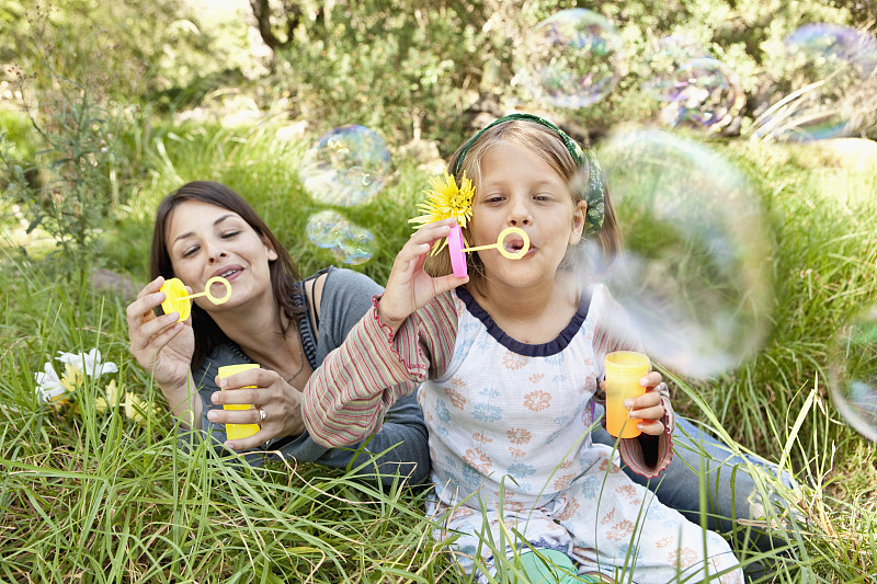 母亲和女儿(10-12)坐在草地上吹泡泡图片素材