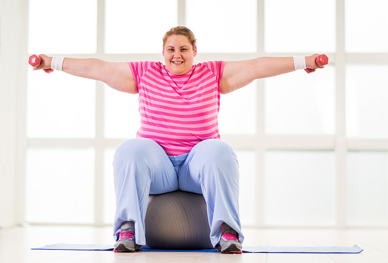 微笑的胖女人坐在健身球和举重。图片素材