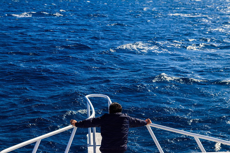 一个人站在游艇的白色船头,看着大海豪华度假的概念后视图图片