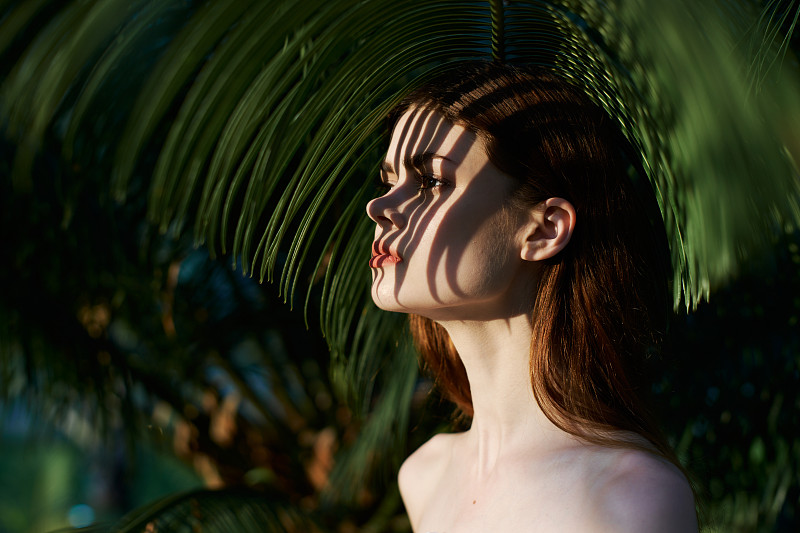 一个年轻美丽的女孩，有着干净健康的皮肤，站在一个热带岛屿上，在棕榈树的树枝的阴影。图片下载