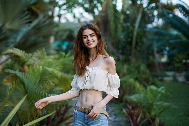 一个穿着超短裤的年轻女子赤裸着肚子在一个热带公园里散步，面带微笑。图片下载