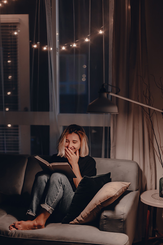 微笑的女人坐在家里的沙发上看书图片素材