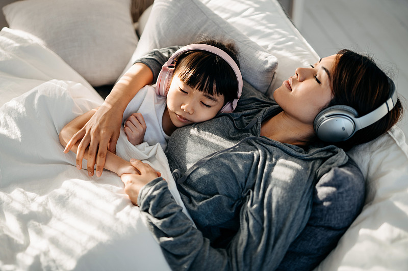 放松的年轻亚洲母亲和小女儿闭着眼睛躺在床上，用耳机听音乐图片素材