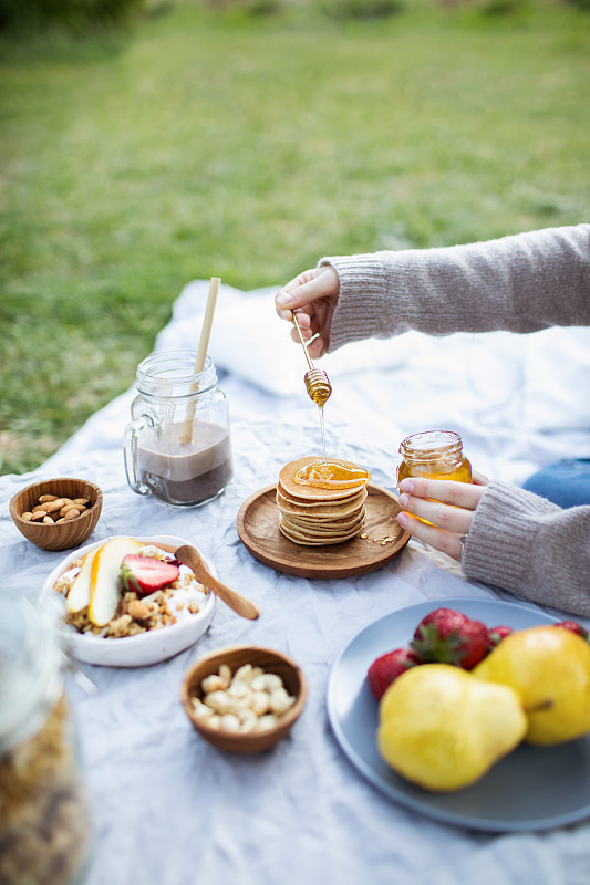 夏季健康素食野餐在公园。
女人用手把有机蜂蜜倒在美味的煎饼上。
水果，可可，蜂蜜煎饼和有机格兰诺拉麦片在亚麻桌布上装饰枕头和柳条篮子。图片素材