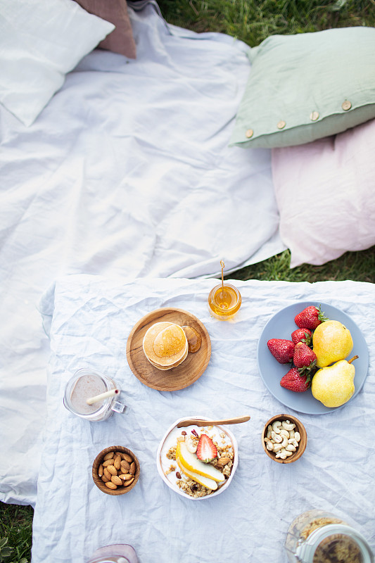夏季健康素食野餐在公园。
水果，可可，蜂蜜煎饼和有机格兰诺拉麦片在亚麻桌布上装饰枕头和柳条篮子。图片下载
