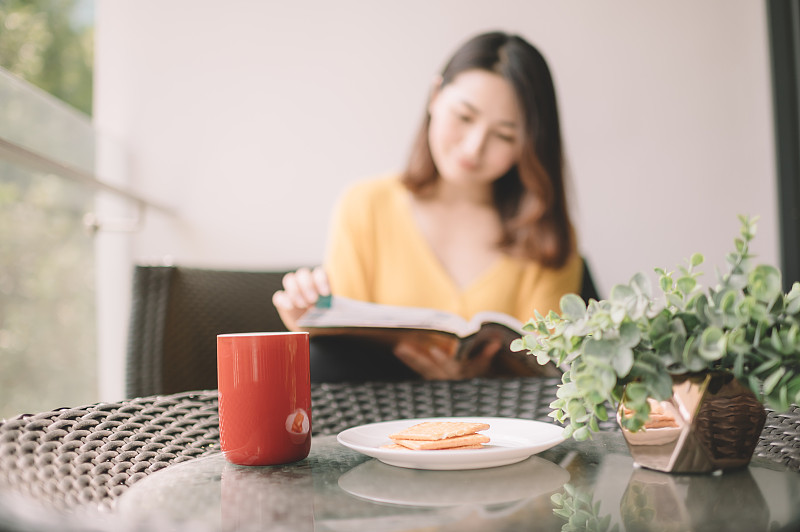 一位亚洲华裔美女中午在她的阳台上看书，放松，享受她的数字排毒时间，喝着咖啡，吃着饼干当点心图片素材