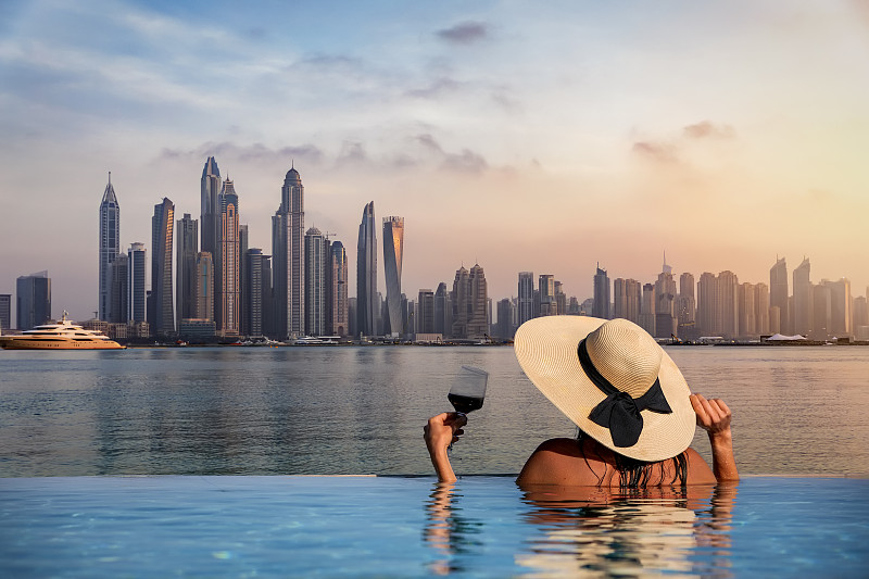 一位戴着帽子的妇女站在泳池边，欣赏着迪拜码头的天际线图片素材