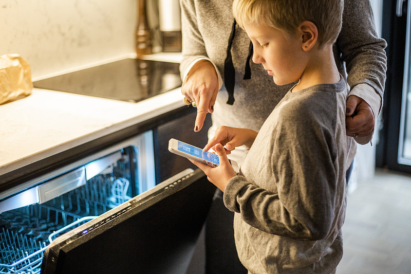 在智能家居的厨房里，男孩通过洗碗机使用手机应用程序，向母亲学习图片素材