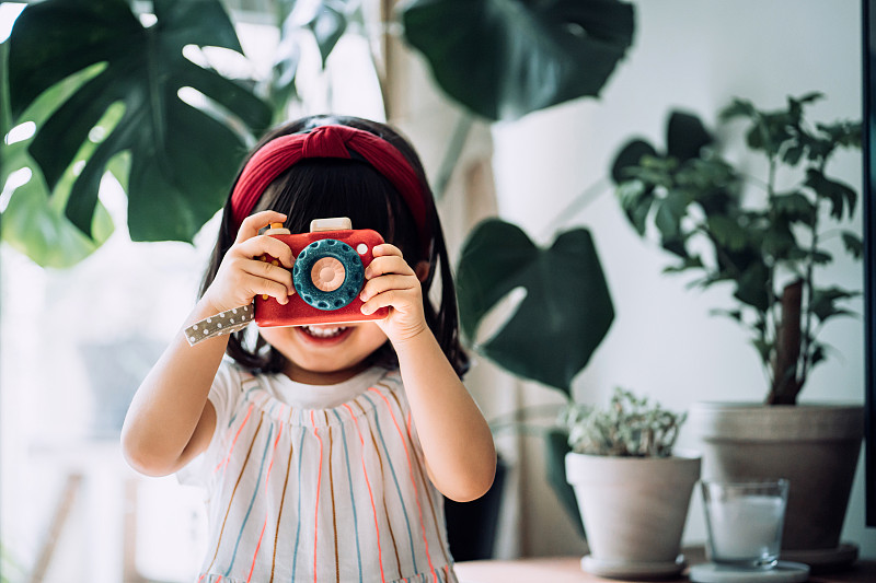 一个带着红头巾的亚洲小女孩，像个专业摄影师一样，用木制玩具相机在家里的盆栽前开心地拍照图片下载