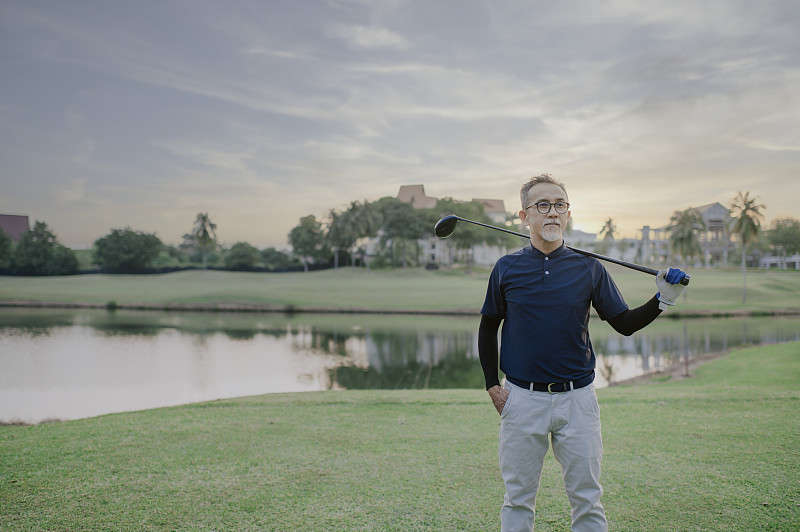 一名亚裔中国资深高尔夫球手肩上扛着高尔夫球杆，看着球场上的景色图片下载