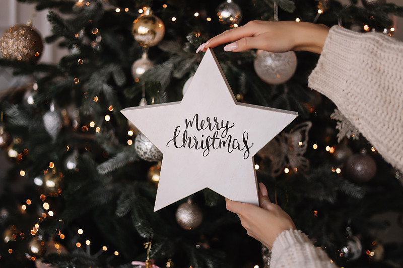在圣诞节和冬天12月的新年期间，一名身穿针织毛衣的妇女手持一颗刻有“圣诞快乐”字样的星星，背景是一棵装饰好的圣诞树图片下载