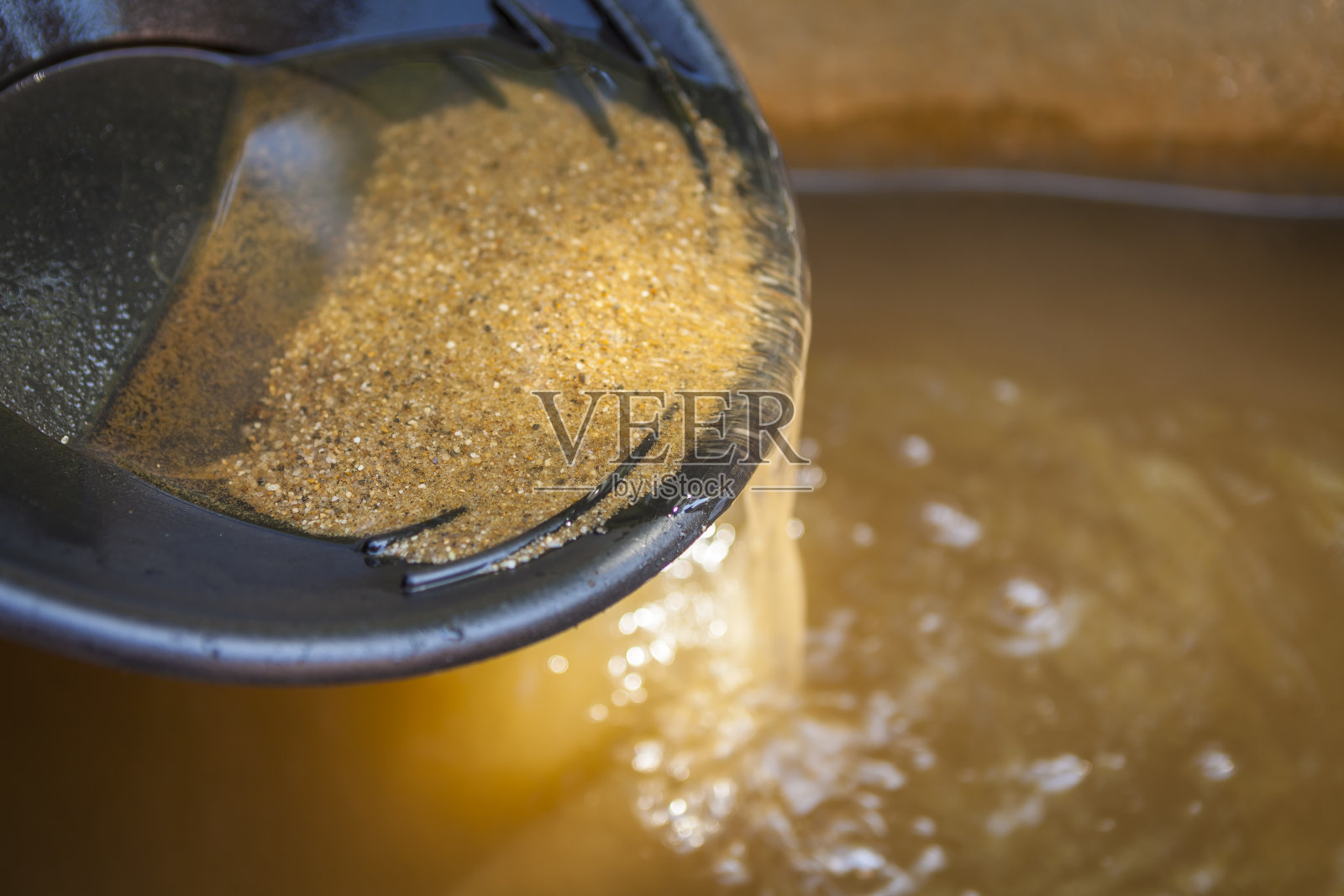 特写的黄金淘金锅与筛砂。景深较浅，聚焦于沙子从锅边流入水中。照片摄影图片