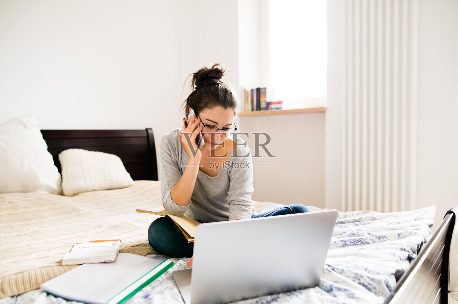 一个女人坐在床上，用笔记本电脑工作，打电话。家庭办公室。照片摄影图片