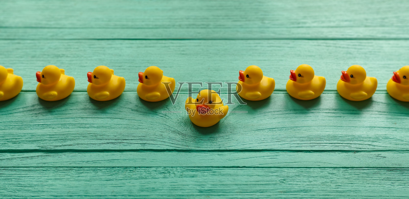 在绿松石色的木桌背景上，一排整齐的黄色橡皮鸭沿着直线移动，一只橡皮鸭从这排橡皮鸭中脱离出来。照片摄影图片