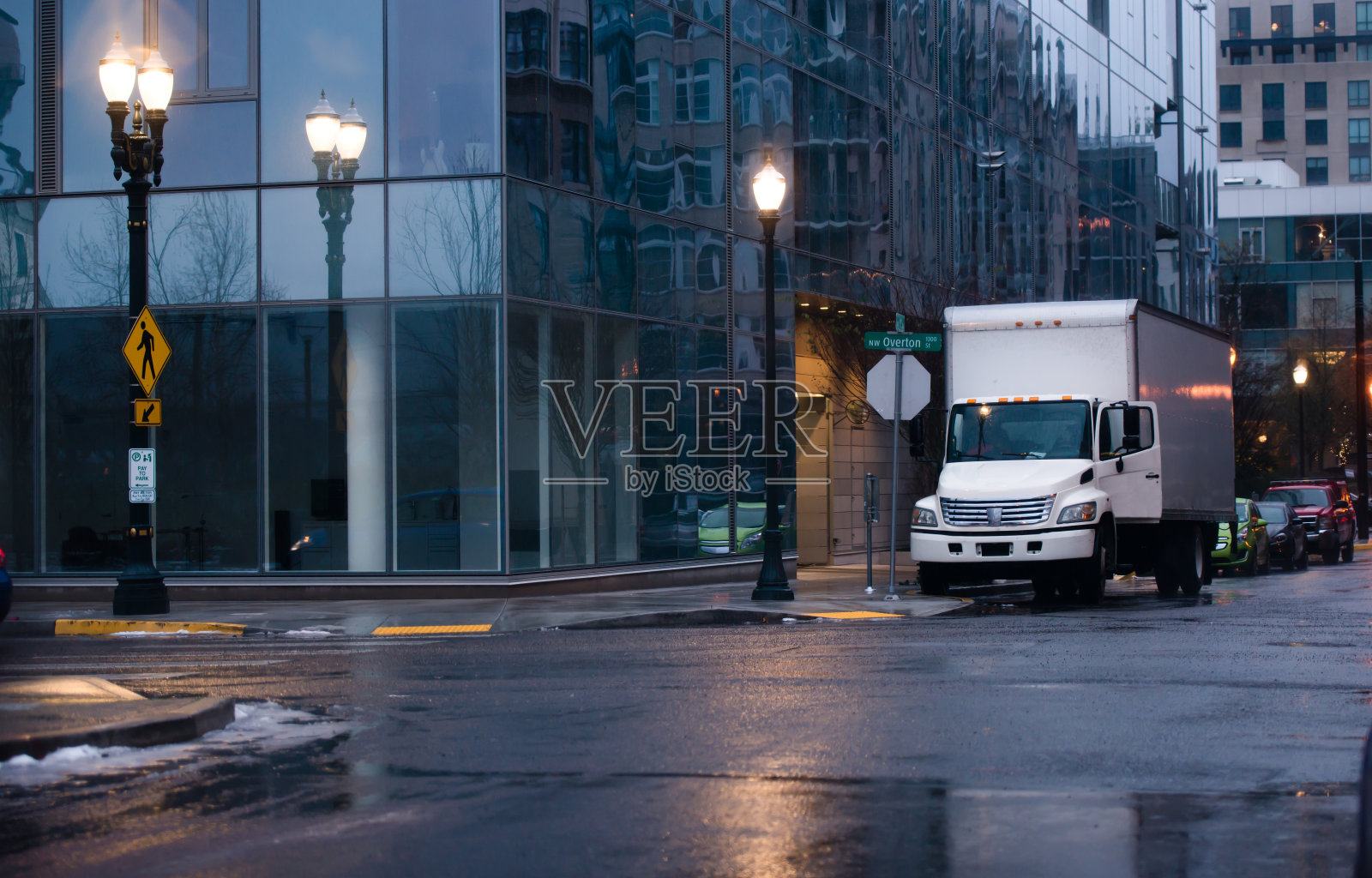 白色小型半挂车与箱式拖车运送货物到现代建筑在夜间城市雨城市街道与光反射。照片摄影图片