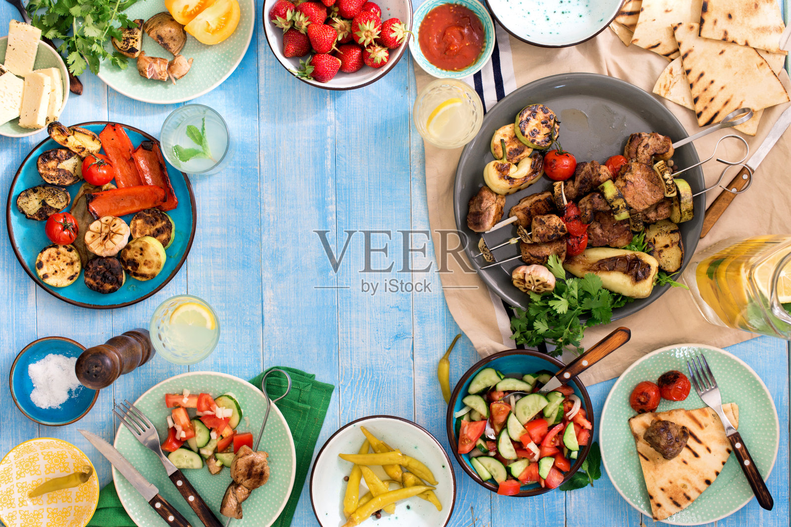 蓝色的木桌上放着烤肉串、烤蔬菜、沙拉、零食、草莓和自制柠檬水照片摄影图片