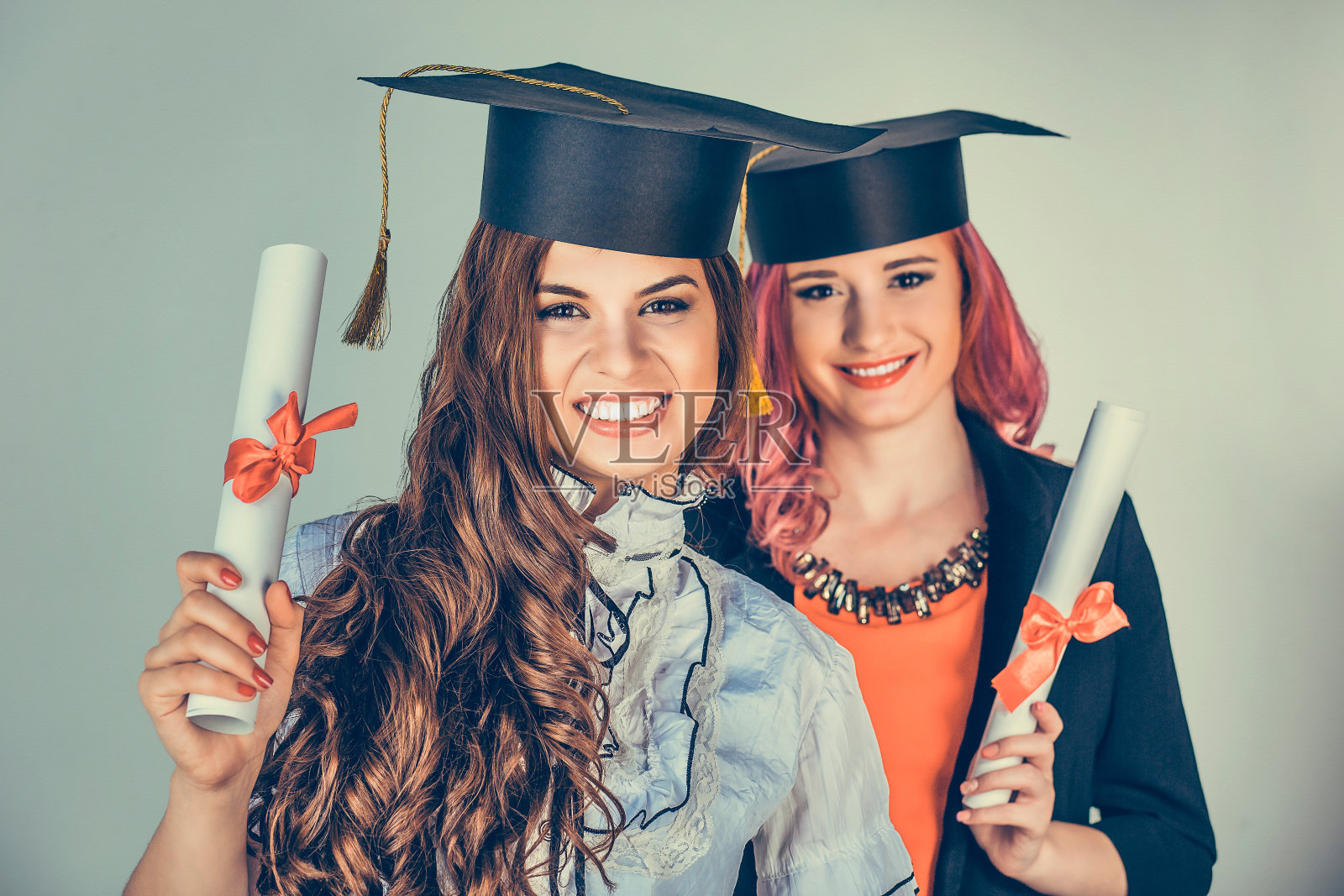 肖像特写美丽快乐的毕业生，两个毕业的女学生，年轻的妇女在帽子长袍转身微笑着拿着文凭卷轴孤立的绿色背景墙。庆祝毕业典礼照片摄影图片