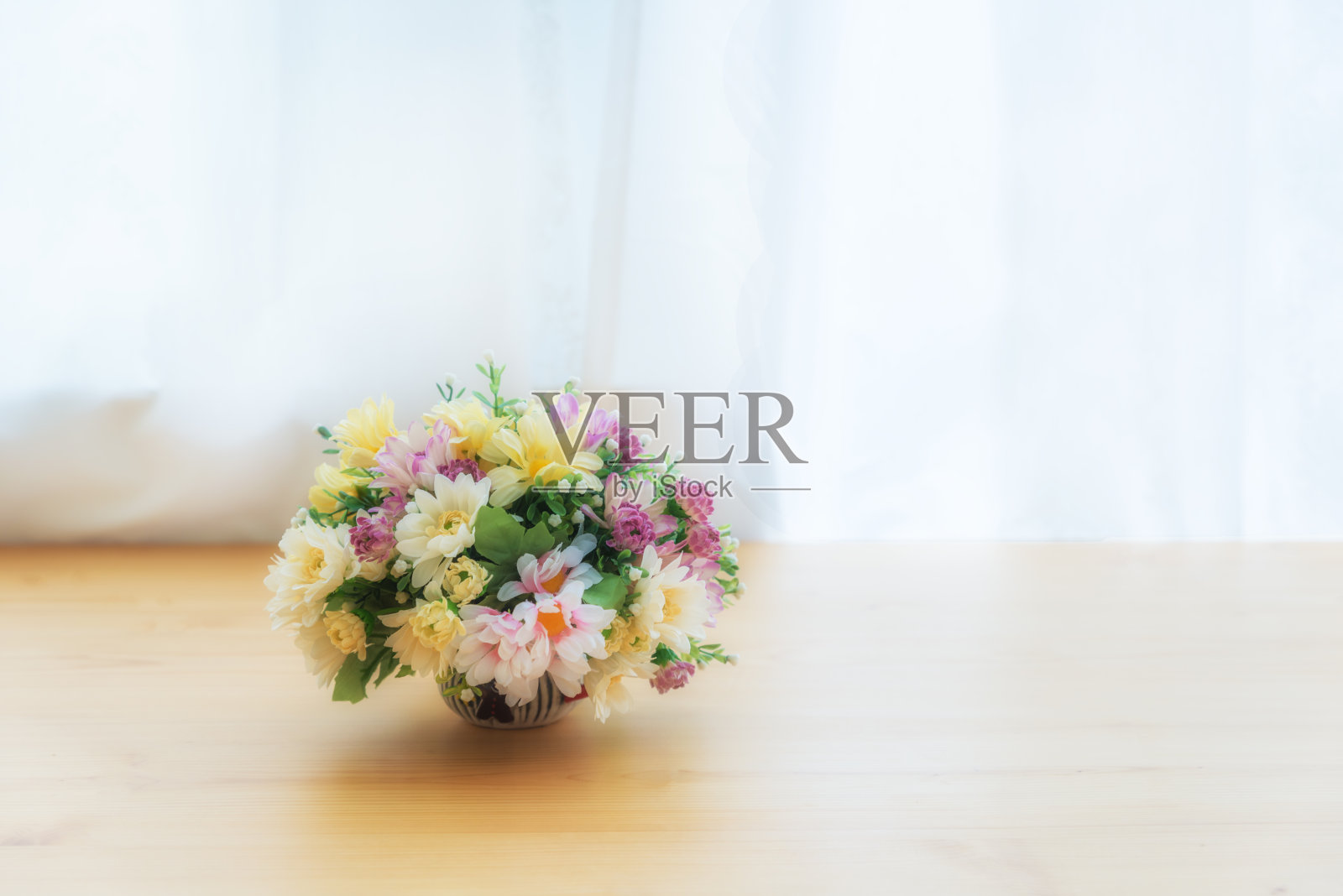 贴着窗帘的木桌上的陶瓷花盆里摆放着五颜六色的人造花。照片摄影图片