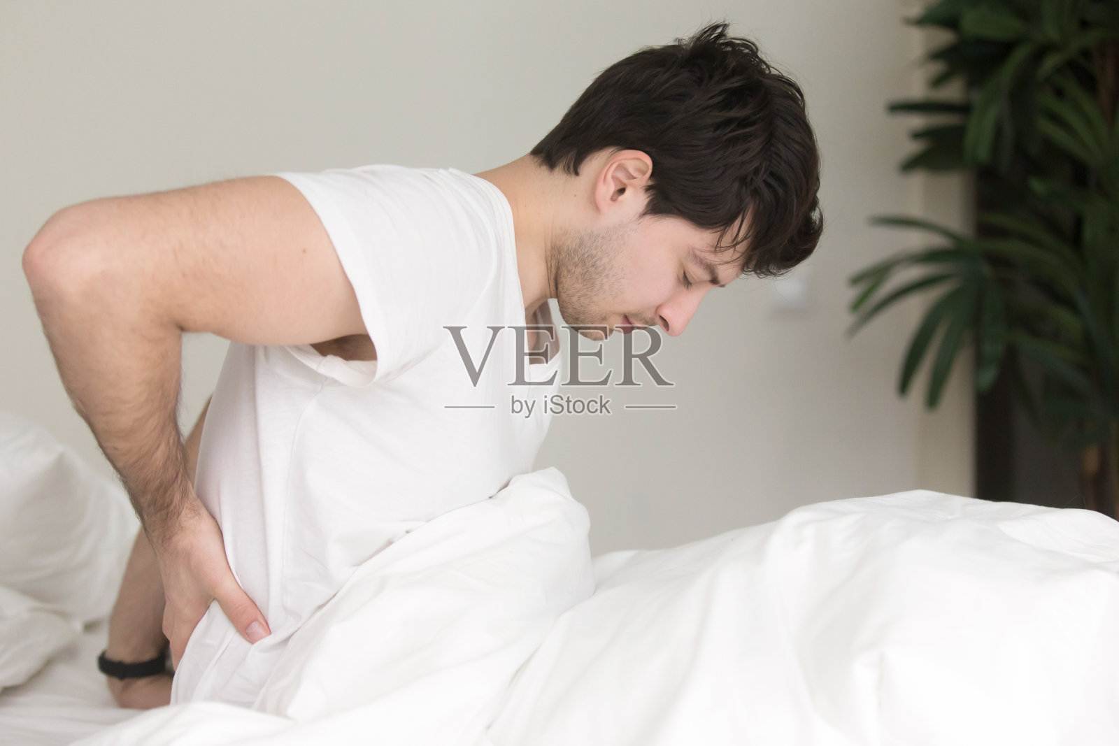 年轻人坐在家里的床上，触摸着疼痛的背部照片摄影图片
