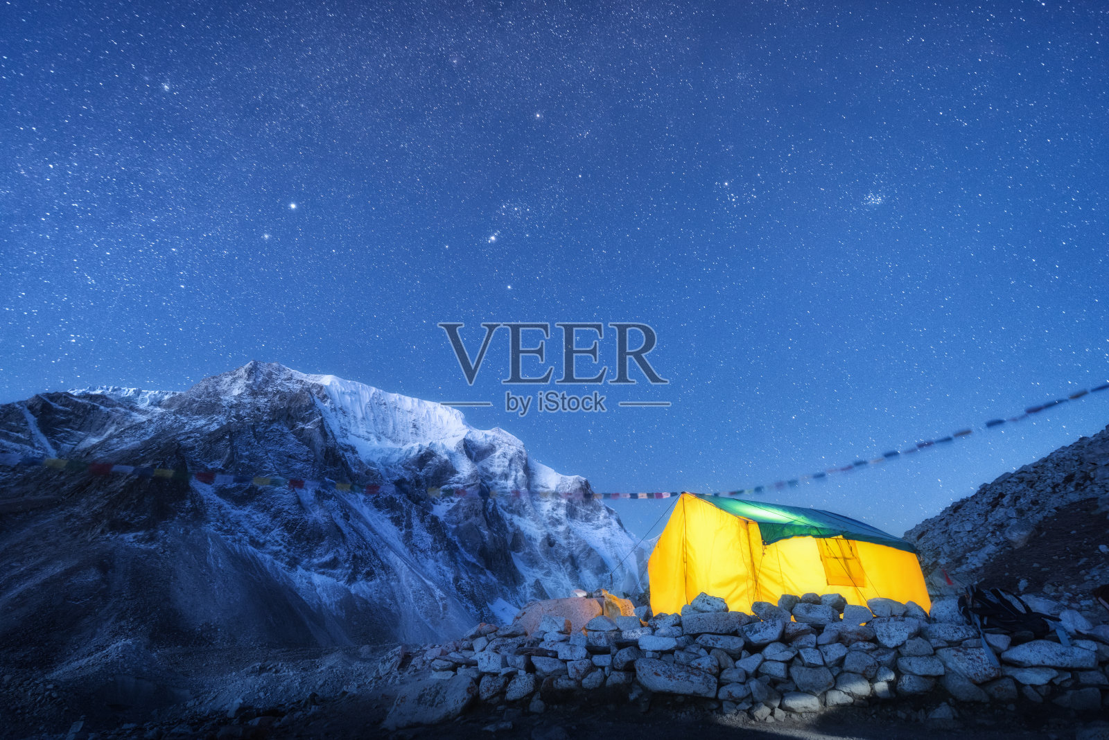 黄色的帐篷，映衬着高高的岩石和白雪皑皑的山峰，夜空中闪烁着星星。喜马拉雅山脉。山川、星空、宿营地。在喜马拉雅山脉徒步旅行。旅行照片摄影图片
