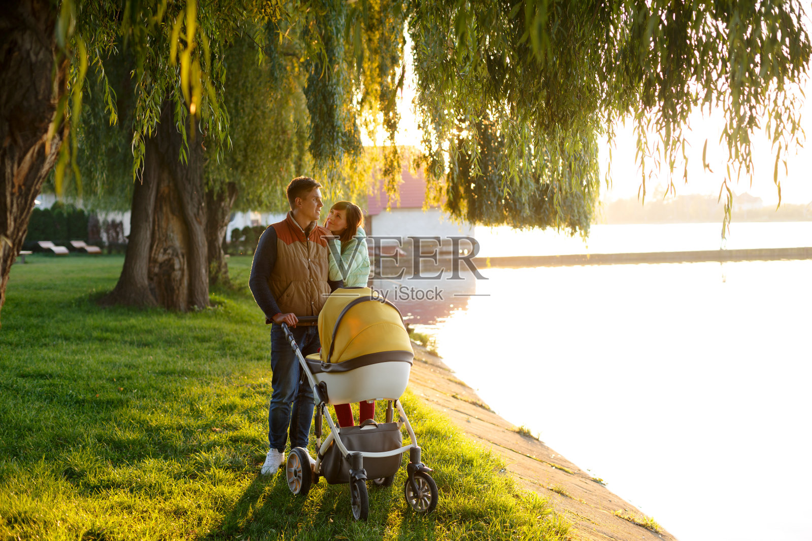一个充满爱的年轻家庭推着婴儿车在湖边散步。微笑的父母夫妇与婴儿婴儿车在秋季公园。爱、亲子、家庭、季节、人的观念。照片摄影图片