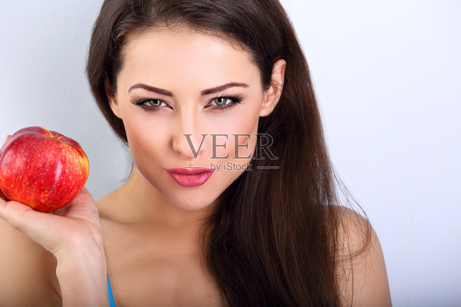 美丽快乐的化妆女人与绿色性感的眼睛持有红色美味的苹果和吹粉红色的嘴唇。特写肖像上的蓝色背景照片摄影图片