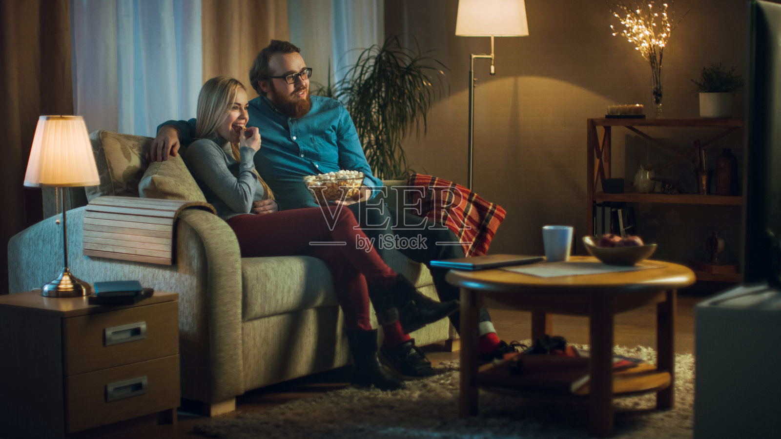 晚上，男人和女人坐在沙发上看电视和吃爆米花。客厅是舒适的。照片摄影图片