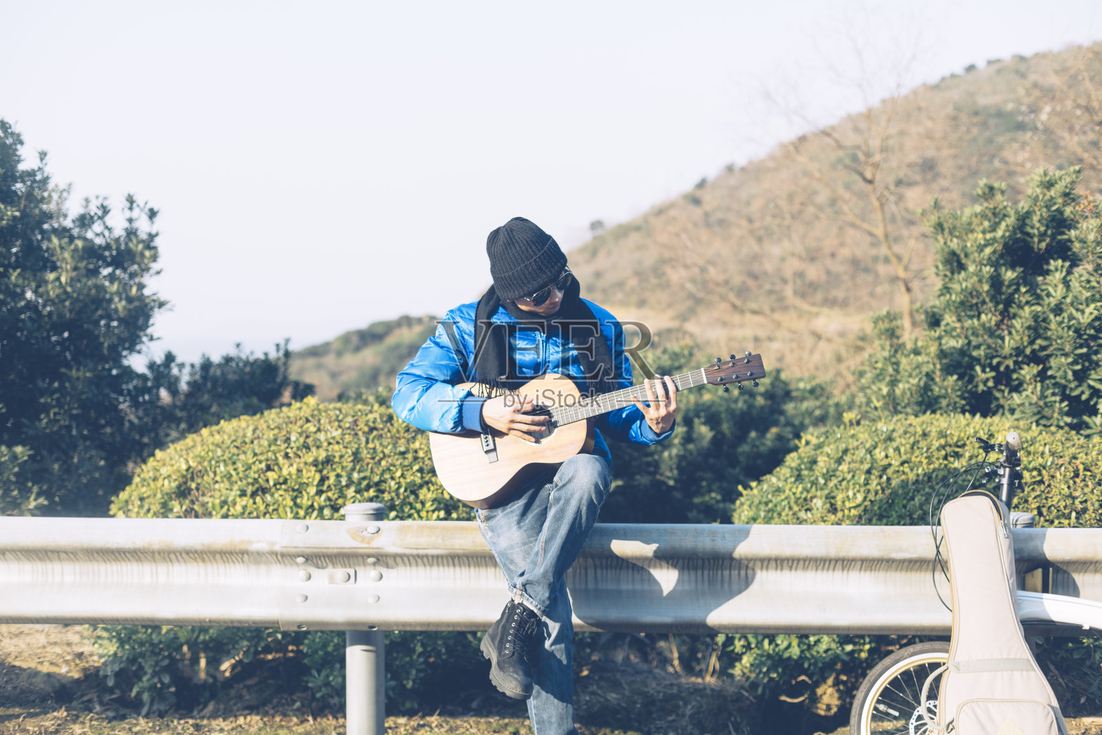 年轻的亚洲男子坐在公路栏杆上弹吉他照片摄影图片
