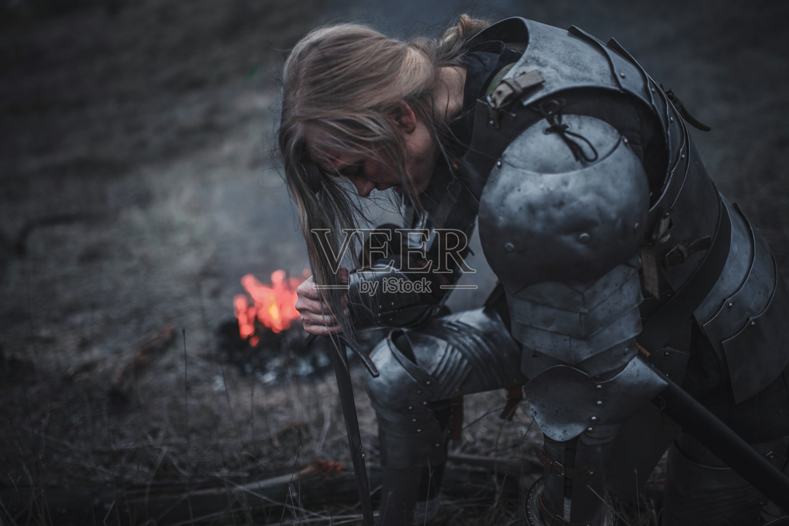 在Jeanne d'Arc的形象中，女孩跪在火和烟的背景下，身穿盔甲，手握宝剑。照片摄影图片