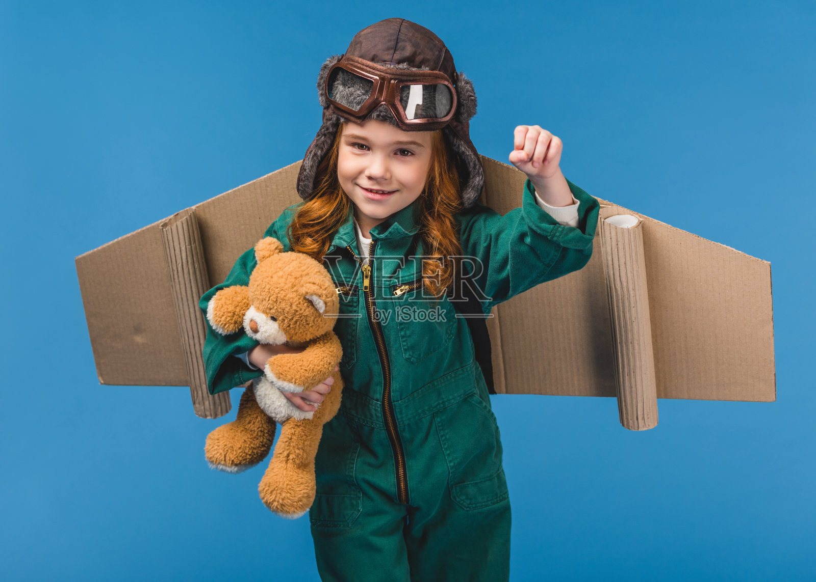 可爱的孩子在飞行员服装与泰迪熊和手工纸飞机机翼孤立在蓝色的肖像照片摄影图片
